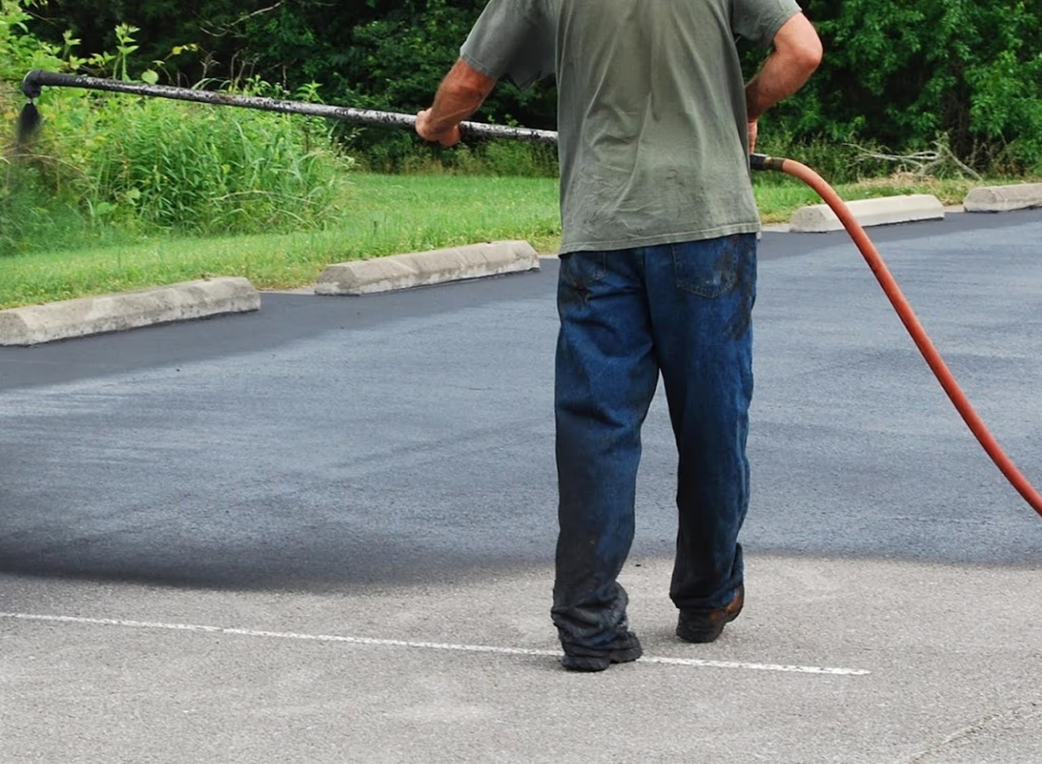 Worker hand-spraying asphalt sealcoat with a red hose along a parking lot edge